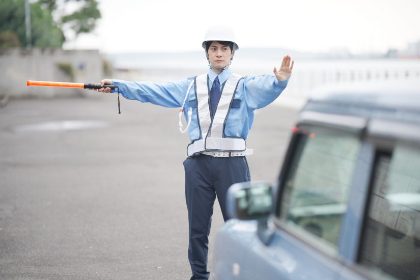 交通整理を行う警備員の男性。ヘルメットと反射ベストを着用し、誘導棒と手で車に停止の合図を送っている。